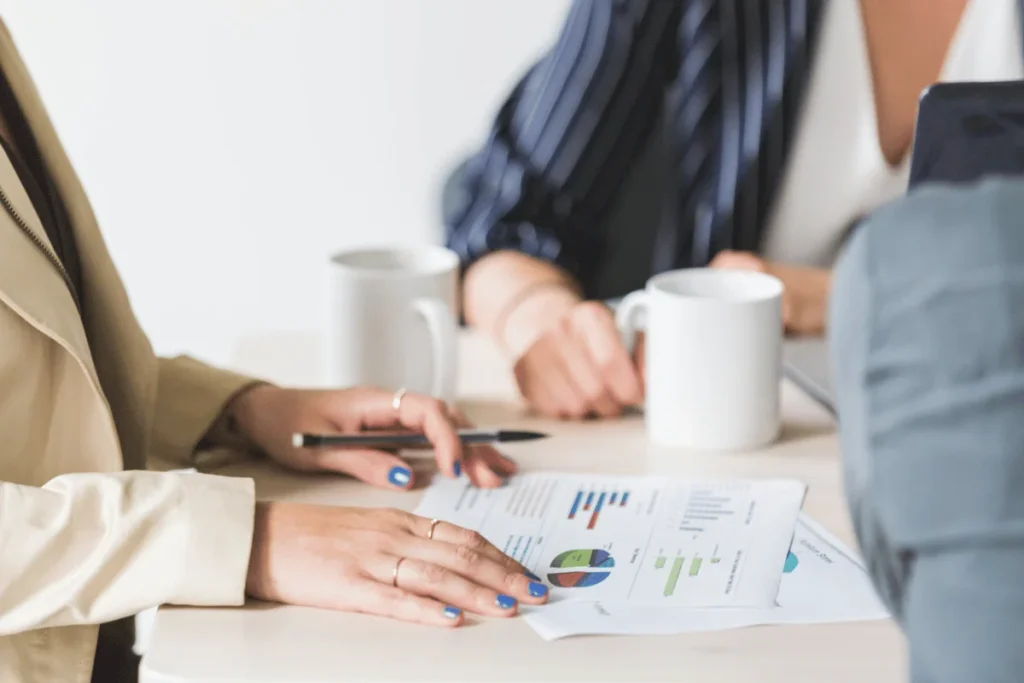 Two people at a table with coffee mugs discuss business charts and graphs, indicating a professional meeting. One has a pen poised for notes.