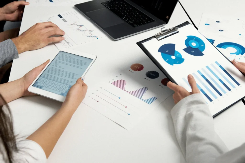 People discussing data with charts on a clipboard, a digital tablet, and printed graphs on a table. The setting is collaborative and focused.