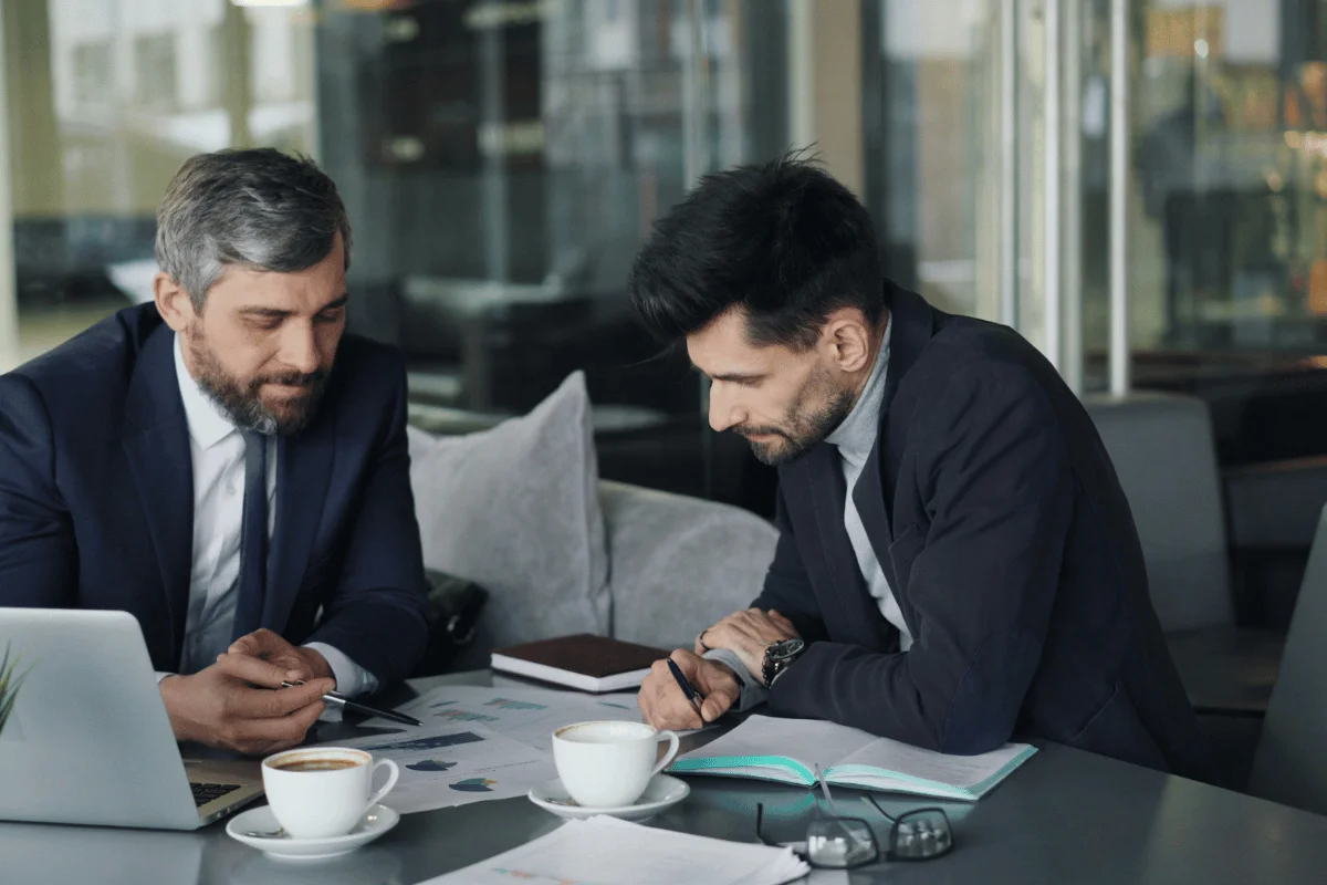 Two businessmen in suits analyzing financial reports with coffee cups.