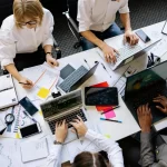 A group of professionals collaborating at a table covered with laptops, documents, and stationery, deep in discussion and work.