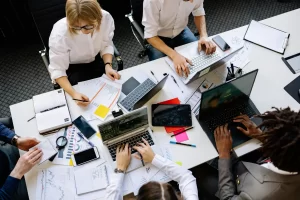 A group of professionals collaborating at a table covered with laptops, documents, and stationery, deep in discussion and work.