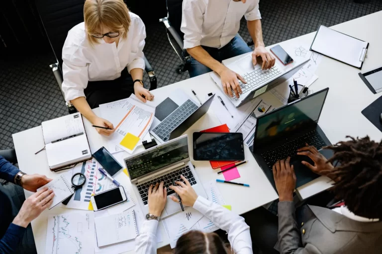 A group of professionals collaborating at a table covered with laptops, documents, and stationery, deep in discussion and work.