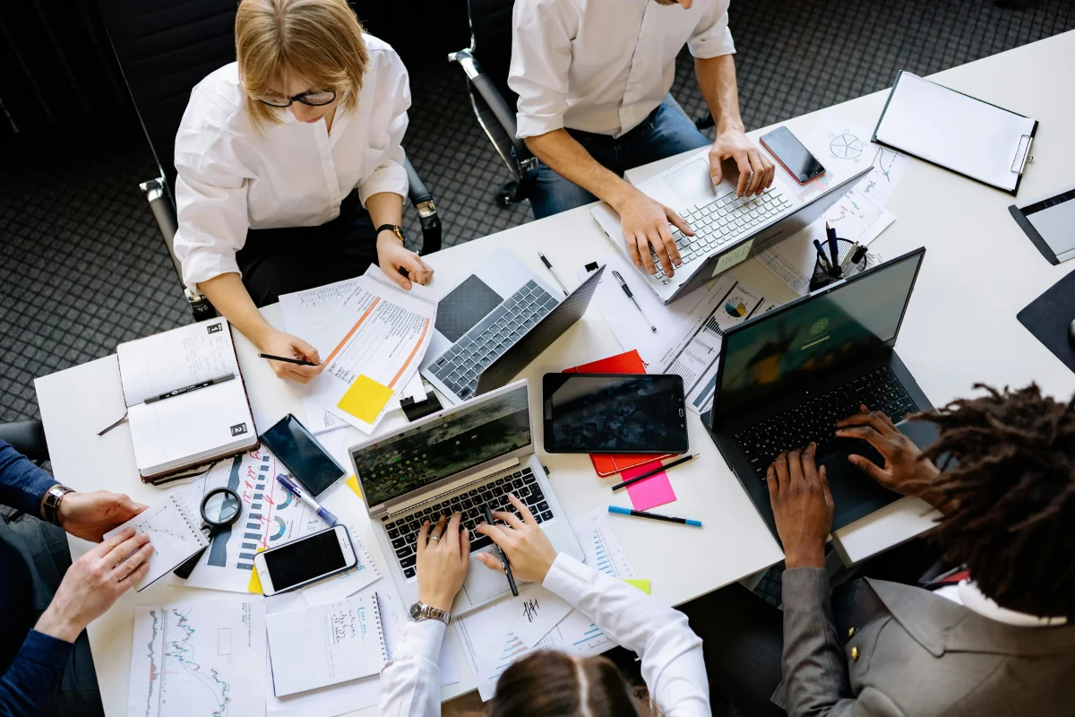 A group of professionals collaborating at a table covered with laptops, documents, and stationery, deep in discussion and work.