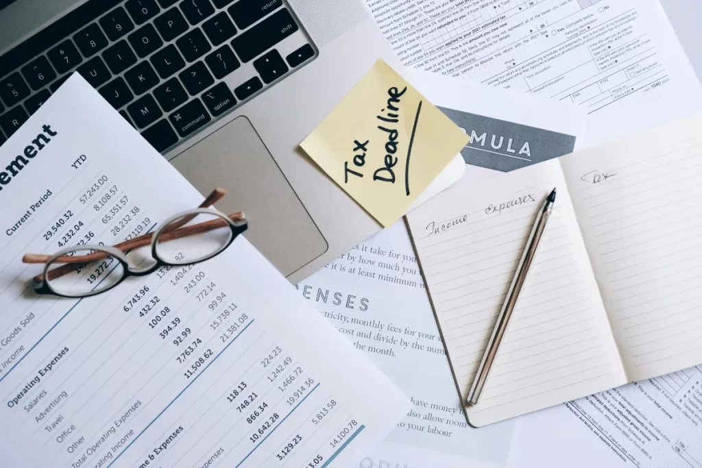A workspace featuring a laptop, financial statements, glasses, and notes with "Tax Deadline" on a sticky note, indicating tax preparation activities.