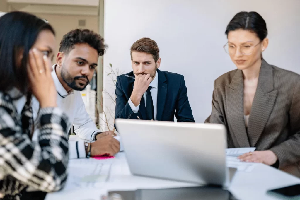 Four professionals in business attire intently focus on a laptop during a meeting. 