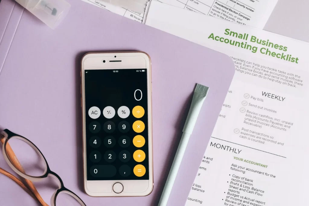 A smartphone with an open calculator app sits on a purple folder next to glasses and a pen. 