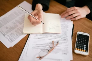 A hand holding a pen writes in a notebook surrounded by papers and a phone displaying a calculator, on a wooden table.