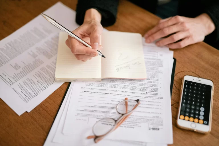 A hand holding a pen writes in a notebook surrounded by papers and a phone displaying a calculator, on a wooden table.