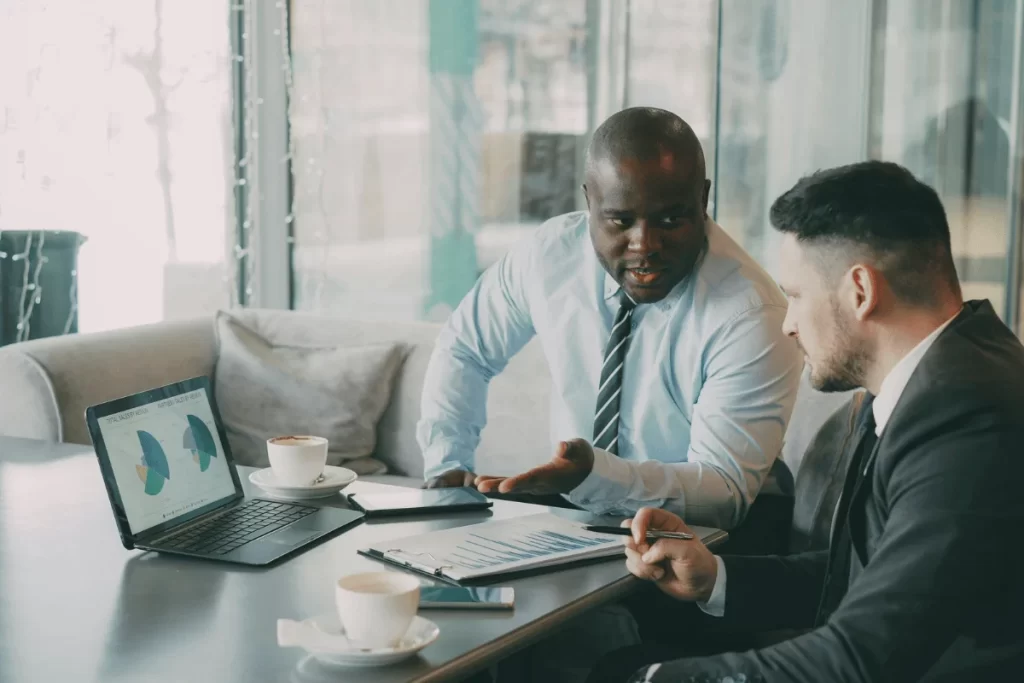 Two men in business attire discuss data at a table with laptops, charts, and coffee. 