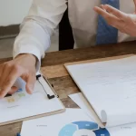 A close-up of a man's hand pointing at a document with graphs and notes on a wooden table during a meeting.