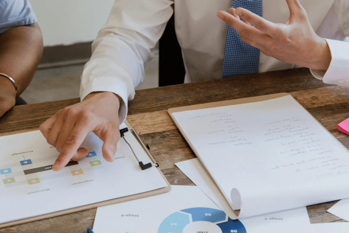 A close-up of a man's hand pointing at a document with graphs and notes on a wooden table during a meeting.