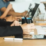 Person holding pen and notebook while collaborating with team in office.