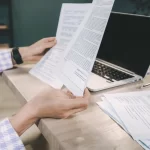 Business professional reviewing financial documents beside a laptop.