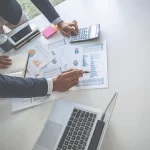 Two professionals in business attire analyze charts and graphs on a desk with a laptop, calculator, and coffee cup, conveying focus and collaboration.