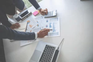 Two professionals in business attire analyze charts and graphs on a desk with a laptop, calculator, and coffee cup, conveying focus and collaboration.