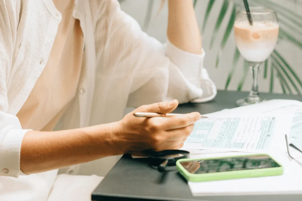Woman in white shirt reviewing paperwork with phone and glasses on table.
