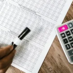 Hand holding pen over tax forms with calculator on wooden desk showing alternative minimum tax Canada calculations.