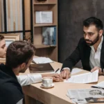 Business consultant reviewing contract documents with a couple during an office meeting at a wooden desk.