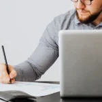 Man in glasses using SaaS valuation calculator on laptop while writing notes at desk.