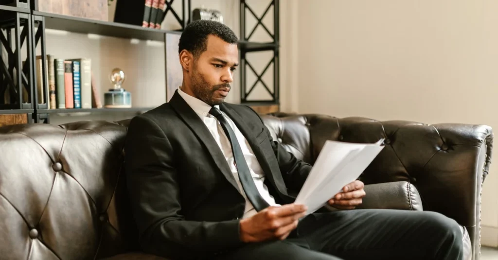 Professional man in business suit reviewing documents on leather couch with shelving behind, sell business Ontario.