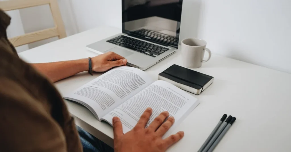 Person studying open book at desk with laptop, phone, coffee, and pens while researching buying existing business.