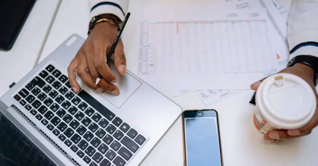Person analyzing buying existing business paperwork with laptop, smartphone, and coffee cup on desk.
