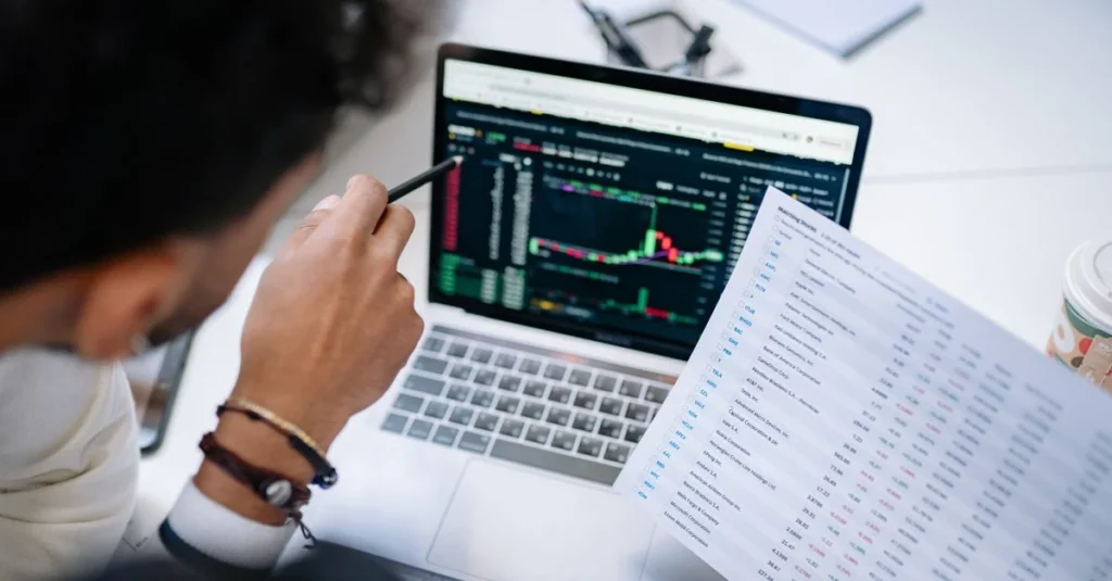 Trader analyzing stock charts on laptop while reviewing financial data spreadsheet.
